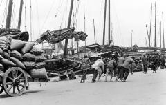 Waterfront showing ship masts and several laborers drawing loaded carts, Hong Kong