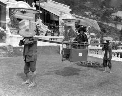 Woman tourist in a sedan chair on poles carried by two workers, Hong Kong